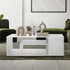 A modern living room with a white coffee table as the central focus, decorated with vases and books. A green sofa is visible in the background. Podgląd zdjęcia produktu