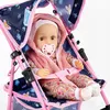 A doll is sitting in a pink and blue patterned doll stroller. Produktbild-Vorschau