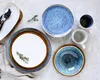 A flat lay of ceramic tableware, including plates, bowls, and mugs, arranged on a light gray surface. Some sprigs of greenery are placed on the plates. Produktbild-Vorschau