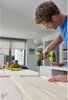 A man is using a cordless drill to work on a wooden surface, with another person in the background. Produktbild-Vorschau
