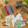 A collection of alcohol markers laid out on a wooden table, with two coloring pages and a hand holding a marker. Podgląd zdjęcia produktu