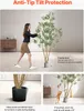 A woman is demonstrating the anti-tip protection of an artificial olive tree, and a dog is shown interacting with it. Voir les images du produit