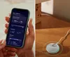 A split image showing a person holding a smartphone displaying a health app and a small white device with a charging cable on a wooden surface. Produktbild-Vorschau