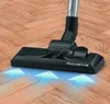 A close-up shot of a vacuum cleaner head on a wooden floor, with blue arrows indicating airflow. Produktbild-Vorschau