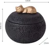 A black, spherical container with a textured surface, topped with a gold-colored cat figurine. Dimensions are indicated by measurement lines and text. Náhled obrázku produktu