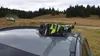 A ski and snowboard carrier is mounted on the roof of a car, with a scenic background of trees and hills. Náhled obrázku produktu