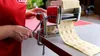 A person is using a red and silver pasta maker to create ravioli. Voir les images du produit