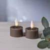 Two LED candles with a taupe base and a white flame-like LED light are placed on a light-colored wooden surface. A blurred background with bokeh lights and a green leaf in the foreground. Produktbild-Vorschau