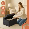 A woman is kneeling on the floor, putting clothes into a dark gray storage ottoman in a living room. Voir les images du produit