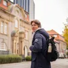 A young man wearing a blue winter coat and a backpack stands on a street in front of a building. Náhled obrázku produktu