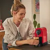 A woman is smiling while holding a cup of coffee next to a red coffee machine. Produktbild-Vorschau