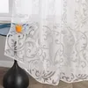 A close-up view of a white sheer curtain with a gray floral pattern, draped over a small table with a glass of orange juice. Produktbild-Vorschau