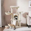 Three cats are interacting with a multi-level cat tree in a living room setting. One cat is lounging in a hanging basket, another is sleeping in a cubby, and a third is eating from a bowl on the floor. Podgląd zdjęcia produktu