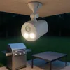 An outdoor motion-activated light fixture mounted under a wooden overhang, illuminating a grill and table. Produktbild-Vorschau
