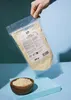 A hand holds a bag of vegan pasta topping over a small wooden bowl filled with the same product, set against a blue background. Produktbild-Vorschau