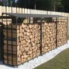 A long, black metal firewood rack filled with stacked logs, situated outdoors next to a wooden fence and a grassy area with white gravel. Podgląd zdjęcia produktu