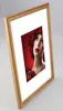 A framed picture of a woman with red hair and makeup, tilted on a light gray surface. Produktbild-Vorschau