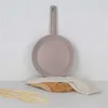 A taupe frying pan is displayed against a textured white wall, resting on a piece of wood with a cloth and some dried wheat stalks. Anteprima dell’immagine prodotto