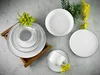 A set of white ceramic plates and cups with black trim, arranged on a textured gray surface, with yellow flowers as decoration. Produktbild-Vorschau