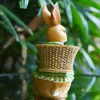A decorative Easter bunny figurine with a basket on its back, set against a blurred green leafy background. Produktbild-Vorschau