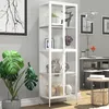 A white glass display cabinet filled with decorative items, placed in a room with a large plant and framed artwork. Podgląd zdjęcia produktu