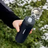 A hand holding a dark blue water bottle with the lid open, outdoors in a snowy and green environment. Náhled obrázku produktu