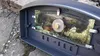 A close-up view of a pizza oven door with a glass window, showing a reflection of trees and foliage. Produktbild-Vorschau