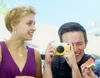 A man and a woman are outdoors, with the man taking a picture with a yellow and white camera. The woman is wearing a purple dress. Náhled obrázku produktu