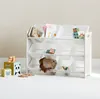 A white toy storage shelf with 12 bins, some containing books and toys, is shown in a room with a light gray wall and white floor. Produktbild-Vorschau