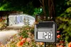 A digital thermometer with a solar panel is placed in a garden at night, with a lit bridge and flower beds in the background. Produktbild-Vorschau