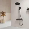 A modern black shower system with a round rain showerhead and a handheld showerhead, mounted on a wall in a bathroom with beige and pink walls and a decorative vase with dried plants on a pedestal. Produktbild-Vorschau