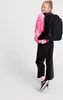A young girl wearing a pink tie-dye sweatshirt and black pants stands with a black backpack on her shoulders, facing away from the camera and looking over her shoulder with a smile. The background is plain white. Produktbild-Vorschau