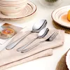 A set of cutlery, including spoons and a fork, laid out on a beige napkin, with plates and a bowl in the background. Produktbild-Vorschau