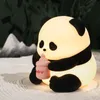 A glowing panda night light sits on a textured surface, holding a pink baby bottle. Produktbild-Vorschau
