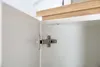 A close-up view of a cabinet hinge on the inside of a white cabinet door. A wooden shelf is visible above. Produktbild-Vorschau