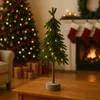 A small, decorated Christmas tree with lights sits on a wooden table in front of a larger, decorated Christmas tree and a fireplace with stockings. Produktbild-Vorschau