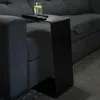 A black C-shaped side table is placed next to a dark gray sofa, with a glass of water and a remote control on top. Produktbild-Vorschau