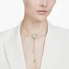 A woman wearing a white blazer and a gold necklace with a flower pendant. Produktbild-Vorschau