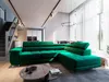 A spacious, modern living room with a large green sectional sofa as the focal point. The room features a staircase, dining area, and large windows with sheer curtains. Produktbild-Vorschau