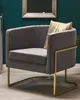 A gray velvet armchair with gold metal legs and a gold striped pillow, set against a teal wall. Náhled obrázku produktu