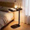 A small side table next to a bed, with a book, cup, and lamp on it. Produktbild-Vorschau