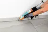 A person wearing gloves is using a caulk gun to apply sealant along the base of a wall where it meets the floor. Produktbild-Vorschau