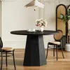 A round black dining table with a vase of pink flowers and two teacups on it, set in a dining room with chairs and a decorative screen in the background. Produktbild-Vorschau