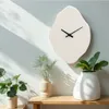 A minimalist wall clock with an organic shape is displayed on a light gray wall, with sunlight casting shadows. Two potted plants in beige and white vases are placed on a wooden shelf in the foreground. Voir les images du produit