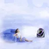 A woman is relaxing on a blue couch in a cloudy, dream-like environment, with a portable fan blowing air towards her. Produktbild-Vorschau