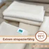 A stack of folded beige cloths, possibly blankets or swaddles, with a label on one of them. Overlay text and a circular badge highlight product features. Produktbild-Vorschau