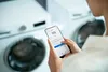 A person is using a smartphone to interact with a washing machine. Produktbild-Vorschau