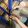 A person is applying a yellow and black striped protective strip to a blue metal rack with double-sided tape. Náhled obrázku produktu