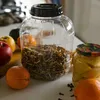 A clear plastic jar filled with dried ingredients, placed on a table with fruits and a jar of pickled items. Produktbild-Vorschau