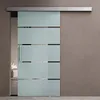 A frosted glass sliding door with horizontal opaque stripes, mounted on a silver track, is shown in a room with a neutral-colored wall and floor. A glimpse of a bedroom interior with a bed and pillows is visible through the partially open door. Produktbild-Vorschau
